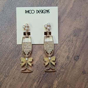May include: A pair of gold tone earrings with a champagne glass design. The glasses are adorned with clear rhinestones and have a yellow bow at the bottom. The earrings are on a white card with the text "Paco Designs".