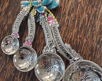 May include: A set of silver measuring spoons with floral designs and embossed measurements. The spoons are connected by a leather cord and adorned with blue and pink decorative accents. Measurements include 1/4 teaspoon, 1/2 teaspoon, and 1 tablespoon.