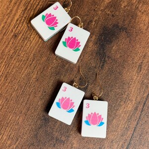 Puede incluir: Dos pares de pendientes rectangulares blancos con forma de fichas de mahjong, cada uno con un gr&aacute;fico de flor de loto rosa y el n&uacute;mero 3 en la esquina superior. Los pendientes cuelgan de ganchos de metal dorado para orejas perforadas.