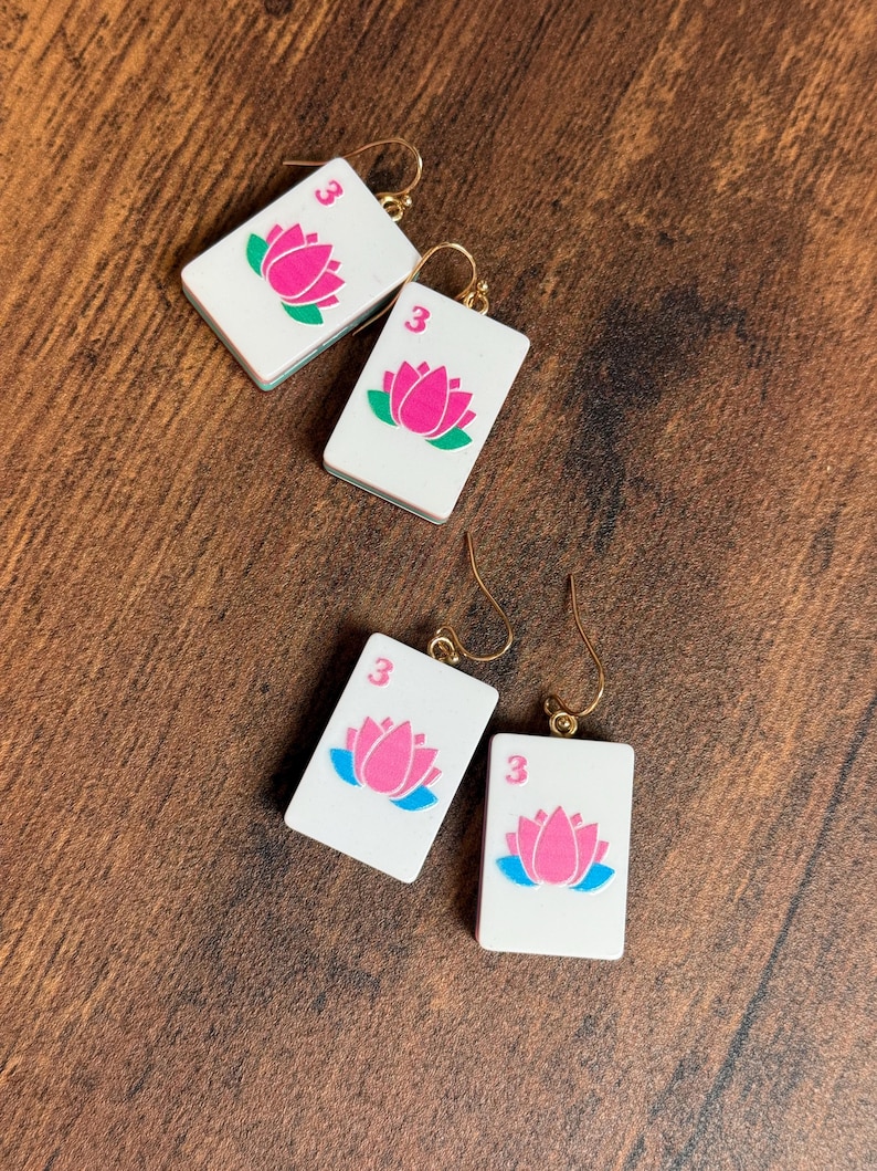 Puede incluir: Dos pares de pendientes rectangulares blancos con forma de fichas de mahjong, cada uno con un gr&aacute;fico de flor de loto rosa y el n&uacute;mero 3 en la esquina superior. Los pendientes cuelgan de ganchos de metal dorado para orejas perforadas.