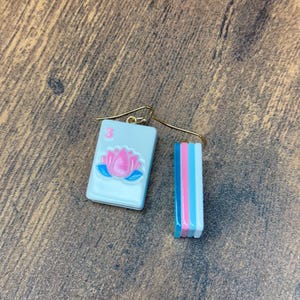Puede incluir: Un par de pendientes colgantes con un dije de ficha de mahjong rectangular con una flor de loto rosa y el n&uacute;mero 3 en rosa. El lado de la ficha muestra rayas verticales en azul, rosa y blanco. Los pendientes tienen ganchos de metal dorado.