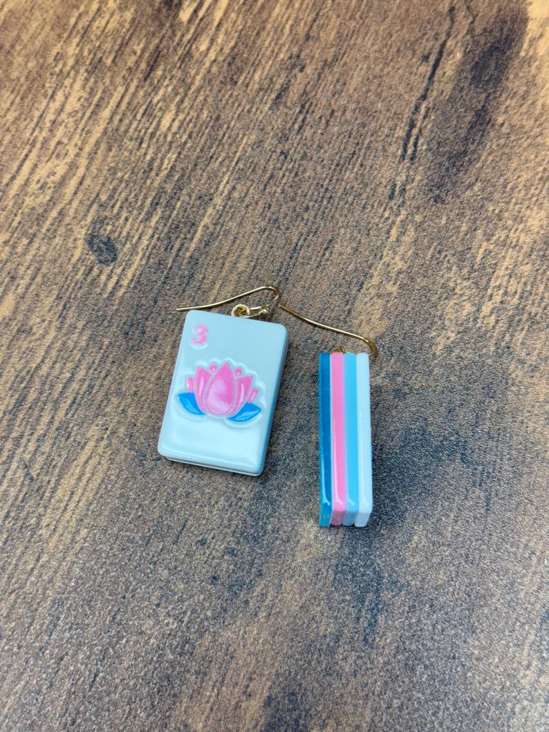 Puede incluir: Un par de pendientes colgantes con un dije de ficha de mahjong rectangular con una flor de loto rosa y el n&uacute;mero 3 en rosa. El lado de la ficha muestra rayas verticales en azul, rosa y blanco. Los pendientes tienen ganchos de metal dorado.