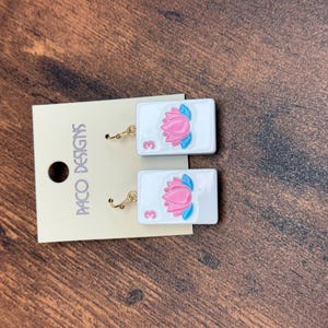 Puede incluir: Un par de pendientes rectangulares blancos con un gr&aacute;fico de flor de loto rosa y el n&uacute;mero 3 en rosa. Los pendientes est&aacute;n sujetos a una tarjeta de presentaci&oacute;n beige con el texto PACO DESIGNS impreso verticalmente en negro a la izquierda.