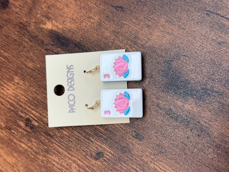 Puede incluir: Un par de pendientes rectangulares blancos con un gr&aacute;fico de flor de loto rosa y el n&uacute;mero 3 en rosa. Los pendientes est&aacute;n sujetos a una tarjeta de presentaci&oacute;n beige con el texto PACO DESIGNS impreso verticalmente en negro a la izquierda.