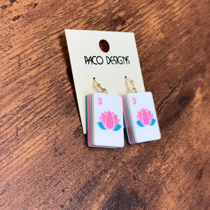Puede incluir: Un par de pendientes colgantes con piezas rectangulares blancas que muestran una flor de loto rosa y el n&uacute;mero 3 en rosa. Las piezas tienen bordes superpuestos en color verde azulado y rosa, suspendidas de ganchos met&aacute;licos dorados.