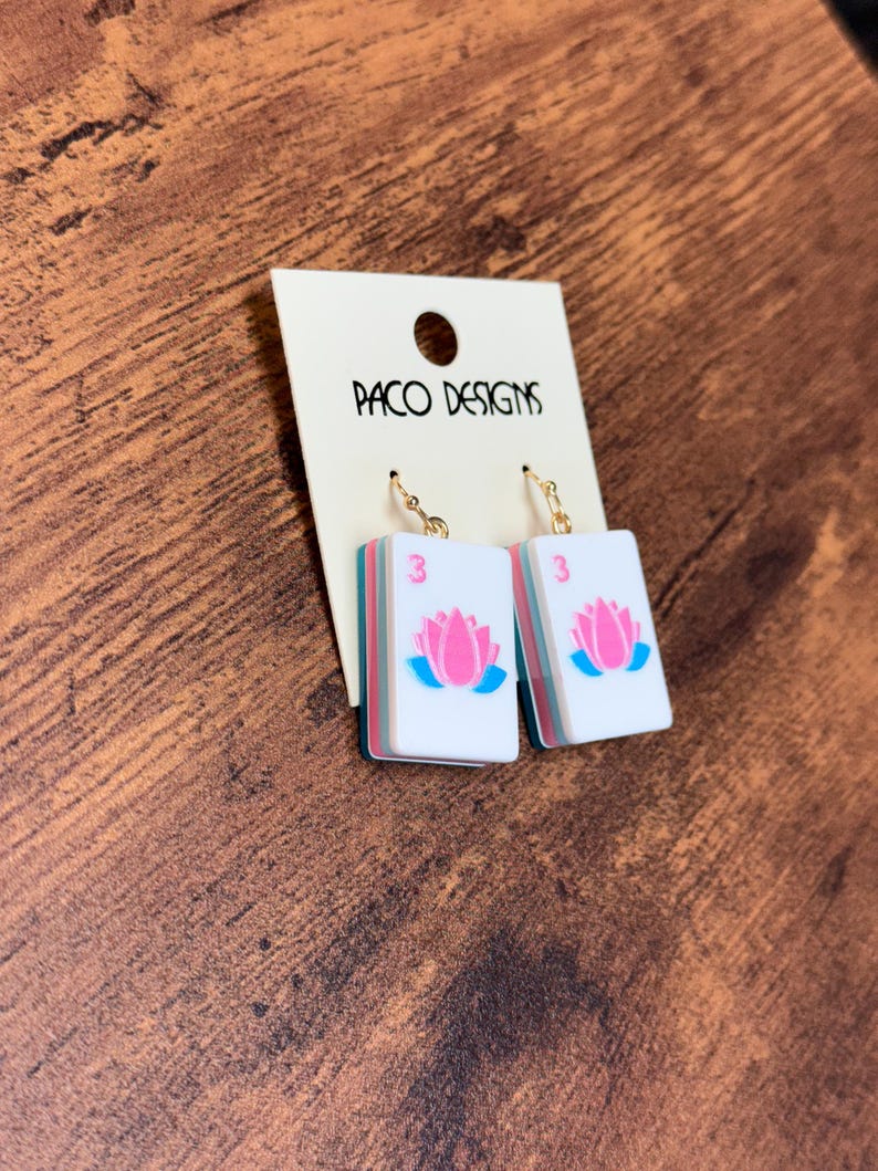 Puede incluir: Un par de pendientes colgantes con piezas rectangulares blancas que muestran una flor de loto rosa y el n&uacute;mero 3 en rosa. Las piezas tienen bordes superpuestos en color verde azulado y rosa, suspendidas de ganchos met&aacute;licos dorados.