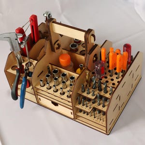 May include: A wooden tool organiser with a handle, featuring multiple compartments for storing various tools, including screwdrivers, drill bits, wrenches, and pliers. The organiser is made of light brown wood and has a natural finish.