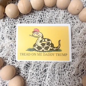 Puede incluir: Una pegatina rectangular con fondo amarillo presenta una serpiente de dibujos animados con gorra rosa y ojos en forma de corazón. La serpiente está enroscada sobre hierba verde. El texto "TREAD ON ME DADDY TRUMP" está impreso debajo.