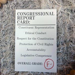 May include: A white Congressional Report Card with black text, including categories like 'Constituent Representation' and 'Ethical Conduct.' The overall grade is an F-, circled in red. The card is surrounded by wooden beads and a white, woven material.