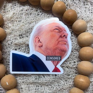 May include: A sticker featuring a portrait of a man with white hair, a blue suit, and a red tie. The word "CENSORED" is printed on a black rectangle over the tie. The sticker is surrounded by wooden beads.