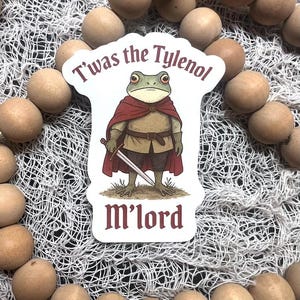 May include: A sticker featuring a cartoon frog dressed as a knight, wearing a red cape and holding a sword. The text "T'was the Tylenol M'lord" is above and below the frog. The sticker is surrounded by light brown spheres and a white net.