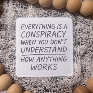 May include: A white square sign with rounded corners features the text "EVERYTHING IS A CONSPIRACY WHEN YOU DON'T UNDERSTAND HOW ANYTHING WORKS" in black. The sign is surrounded by a white mesh and wooden beads.