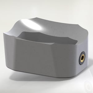 Op de afbeelding: Een grijs, rechthoekig object met een gebogen bovenkant en een verzonken gebied. Het object heeft een kleine, cirkelvormige opening met een goudkleurige ring aan één kant. Het ontwerp is modern en minimalistisch.