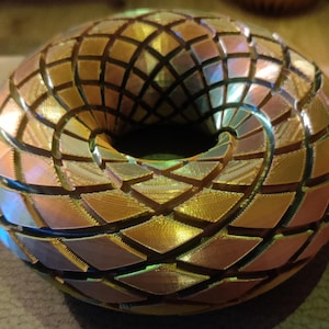 Pode incluir: Um objeto geométrico impresso em 3D com a forma de um donut. O objeto é feito de um material brilhante e iridescente com um esquema de cores dourado e cobre. A superfície do objeto é coberta por uma grade.