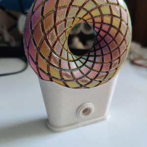 Può includere: Una sfera iridescente stampata in 3D con un foro al centro, che poggia su una base bianca. La sfera presenta un motivo geometrico e sembra essere fatta di plastica.