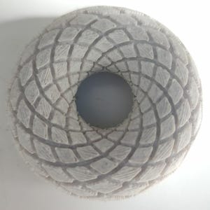 Könnte beinhalten: Eine weiße, donut-förmige Skulptur mit strukturierter Oberfläche. Das Design zeigt ein sich wiederholendes Rautenmuster, das sich um das zentrale Loch windet. Die Skulptur besteht aus einem festen, undurchsichtigen Material.