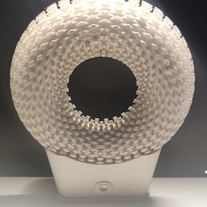 Könnte beinhalten: Eine weiße, 3D-gedruckte, ringförmige Lampe mit einer strukturierten Oberfläche. Die Lampe hat einen kleinen Sockel und ein Loch in der Mitte.