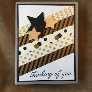 May include: A white card with a gray border features a diagonal design of black and gold washi tape. The washi tape has different patterns, including stripes, polka dots, and zigzags. Three gold and black stars are layered on top of the tape. The card reads "Thinking of you".
