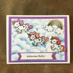 May include: A purple card with a welcome baby message and three unicorns flying in a blue sky with clouds and a rainbow.