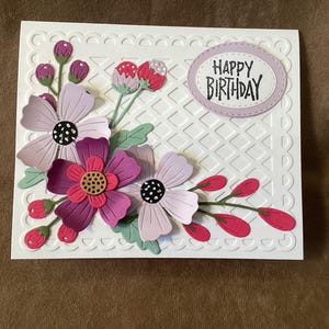 May include: A white birthday card with a floral design. The card features layered paper flowers in shades of purple, pink, and green. The words "HAPPY BIRTHDAY" are in a light purple oval. The card has a decorative border.