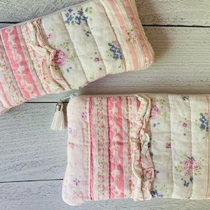 May include: Two rectangular quilted pouches featuring a patchwork design of pink and white stripes, floral patterns, and a ruffled fabric accent. Each pouch includes a white zipper closure with a small white tassel attached to the zipper pull.