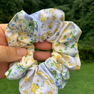May include: A floral scrunchie with a mix of white, yellow, and blue fabric. The scrunchie features a delicate floral pattern, with small yellow and blue flowers on a white background. The accessory is held up against a green background.