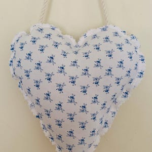 May include: A heart-shaped wall hanging with a white background and a blue floral pattern. The edges are scalloped and trimmed with white fringe. A white rope is attached for hanging.