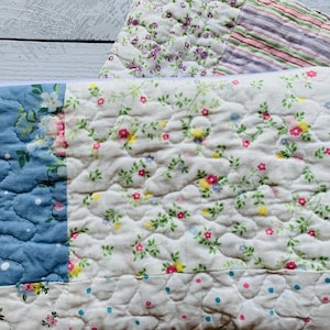 May include: A rectangular quilted pouch featuring a patchwork design with a blue floral section, a white section with small pink and yellow flowers, and a striped section with pink, green, and purple lines. The pouch has a white zipper closure.