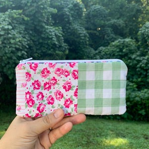 May include: A rectangular, quilted pouch with a white zipper. The pouch is divided into three sections: a floral print with pink roses, a green and white gingham pattern, and a striped section. The pouch is held in a hand.