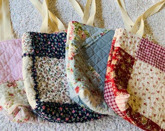 May include: Four quilted tote bags with cream-colored fabric handles. The bags feature various patchwork designs including light pink, navy blue with small flowers, light blue with floral patterns, and red with white checkered and floral prints.