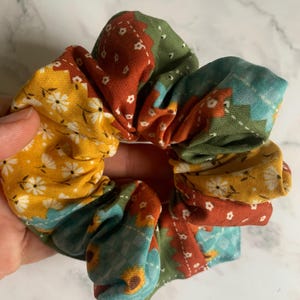 May include: A fabric hair scrunchie featuring a patchwork design with various floral and geometric patterns in shades of mustard yellow, rust orange, sage green, and light blue. The accessory has a gathered, ruffled texture throughout the fabric.