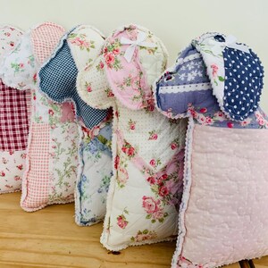 May include: A row of handmade dog-shaped pillows in various floral and patterned fabrics. The pillows feature a patchwork design with colors including red, pink, blue, and white. Each pillow has a unique fabric combination and design.