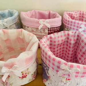 May include: Five fabric storage baskets in various patterns and colors. The baskets feature floral, gingham, and patchwork designs in shades of pink, blue, and white. Each basket has a contrasting interior and is tied with a white bow.