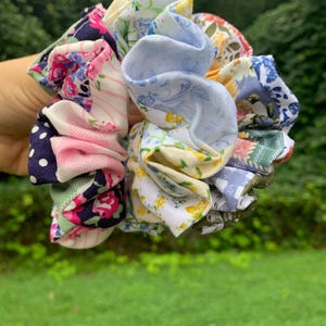 May include: A fabric hair scrunchie featuring a patchwork design of various floral, polka dot, and striped patterns in shades of pink, blue, yellow, green, and navy. The accessory has a gathered, ruffled texture created by an internal elastic band.