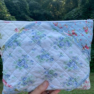 May include: A quilted pouch with a floral pattern in shades of blue, pink, and white. The pouch has a zipper closure and a diamond-stitched design. The edges are trimmed with ruffled fabric.