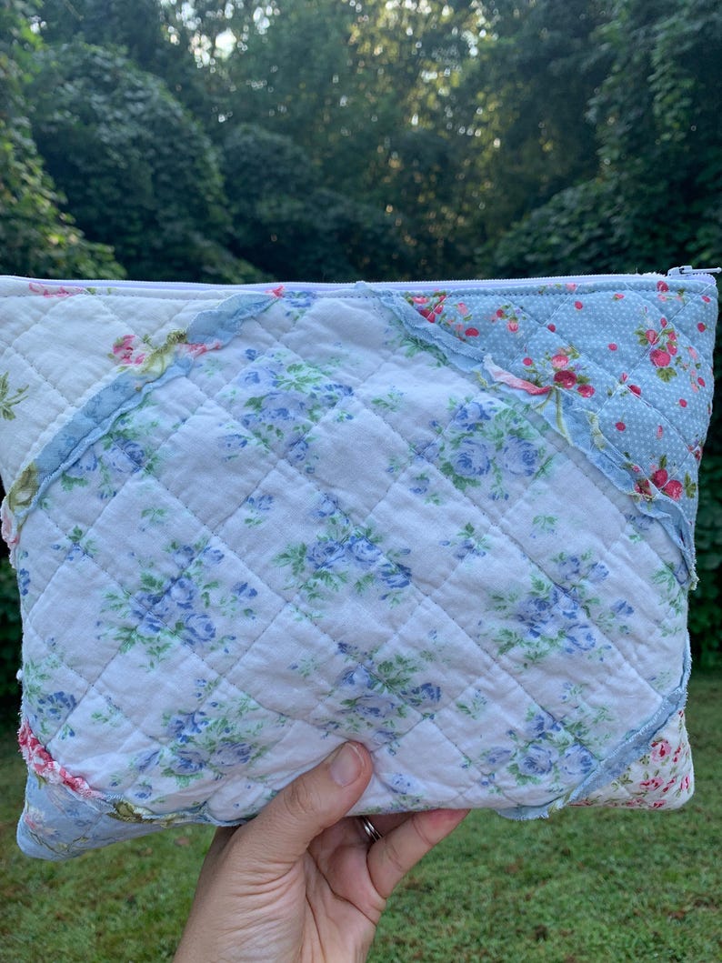 May include: A quilted pouch with a floral pattern in shades of blue, pink, and white. The pouch has a zipper closure and a diamond-stitched design. The edges are trimmed with ruffled fabric.