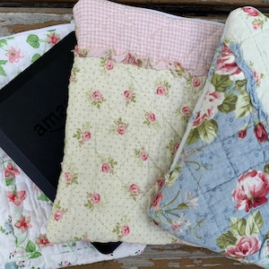 May include: A collection of quilted fabric items with floral patterns. The items include a white quilt with pink and blue flowers, a light blue quilt with pink roses, and a cream-colored quilt with pink roses and green leaves. A black tablet is partially visible.