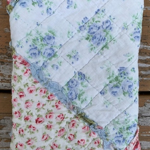 May include: A quilted fabric item with a floral pattern. The top section features blue roses on a white background, while the bottom section has pink roses. The edges are trimmed with ruffled fabric in blue and pink.