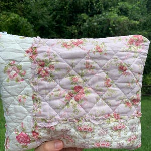 May include: A quilted pillow with a floral pattern in shades of pink, white, and green. The pillow features a diamond pattern and ruffled edges. The design includes roses and floral basket motifs, creating a vintage, cottagecore aesthetic.
