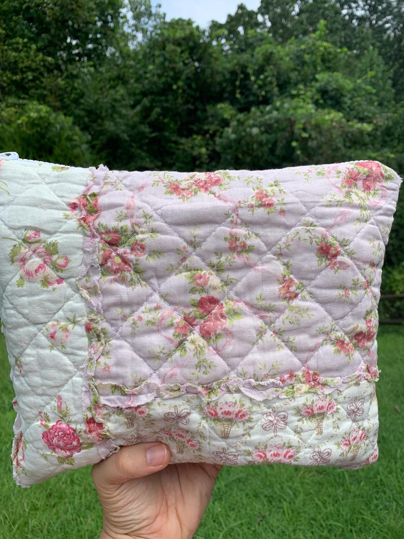 May include: A quilted pillow with a floral pattern in shades of pink, white, and green. The pillow features a diamond pattern and ruffled edges. The design includes roses and floral basket motifs, creating a vintage, cottagecore aesthetic.