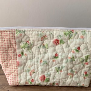 May include: A quilted cosmetic bag with a floral pattern and a pink gingham accent. The bag features a white zipper and a pink tassel. The floral pattern includes pink roses and green leaves on a cream background. The bag is on a wooden surface.
