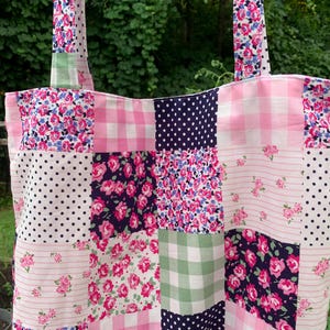 May include: A patchwork tote bag with a variety of floral, polka dot, and gingham patterns in pink, white, and navy blue. The bag has two long straps and is made of fabric. The bag is designed for carrying items.