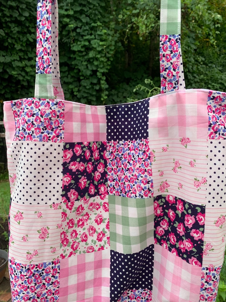 May include: A patchwork tote bag with a variety of floral, polka dot, and gingham patterns in pink, white, and navy blue. The bag has two long straps and is made of fabric. The bag is designed for carrying items.