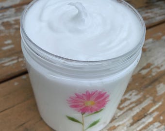 May include: A clear plastic jar filled with white cream. The jar has a pink flower design on the side. The cream has a swirl pattern on top, suggesting a whipped or textured consistency. The jar is sitting on a wooden surface.