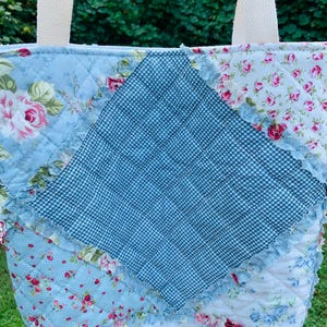 May include: A quilted tote bag featuring a patchwork design with a central dark blue and white gingham diamond panel. The bag is constructed from various fabric squares displaying light blue floral patterns, pink roses, and ruffled seams.