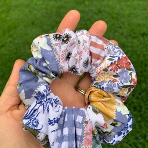 May include: A colorful scrunchie with a patchwork design. The fabric features floral and geometric patterns in shades of blue, white, yellow, red, and green. The scrunchie is held in a hand against a blurred green background.
