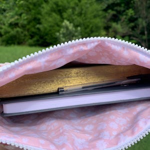 May include: A pink floral-patterned pouch with a white zipper, holding a gold-covered book, a gray notebook, and a pen. The pouch's interior features a delicate white floral design on a pink background, suggesting a feminine aesthetic.