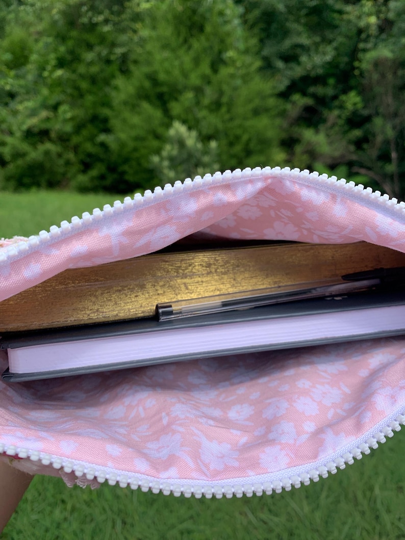 May include: A pink floral-patterned pouch with a white zipper, holding a gold-covered book, a gray notebook, and a pen. The pouch's interior features a delicate white floral design on a pink background, suggesting a feminine aesthetic.