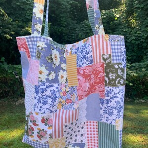 May include: A patchwork tote bag with floral, striped, and gingham patterns in various colors. The bag has long straps and is made of fabric. The background is a blurred view of green foliage.