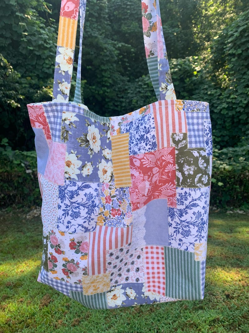 May include: A patchwork tote bag with floral, striped, and gingham patterns in various colors. The bag has long straps and is made of fabric. The background is a blurred view of green foliage.